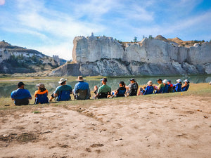 People sitting together on the bank of a lake overlooking a beautiful sky and mountains