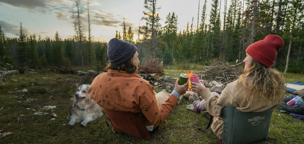 Two people cheersing their drinks while sitting in crazy creek chairs in front of a bonfire next to their dog