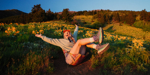 Person sitting in a flower printed Crazy Creek hex 2.0 chair with arms outstretched in a field of yellow flowers at sunset