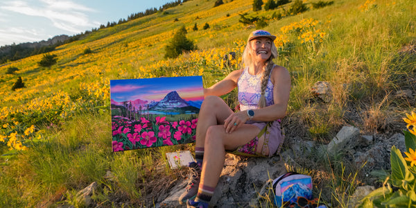 Rachel Pohl sitting in a field of flowers holding a painting of a mountain landscape.