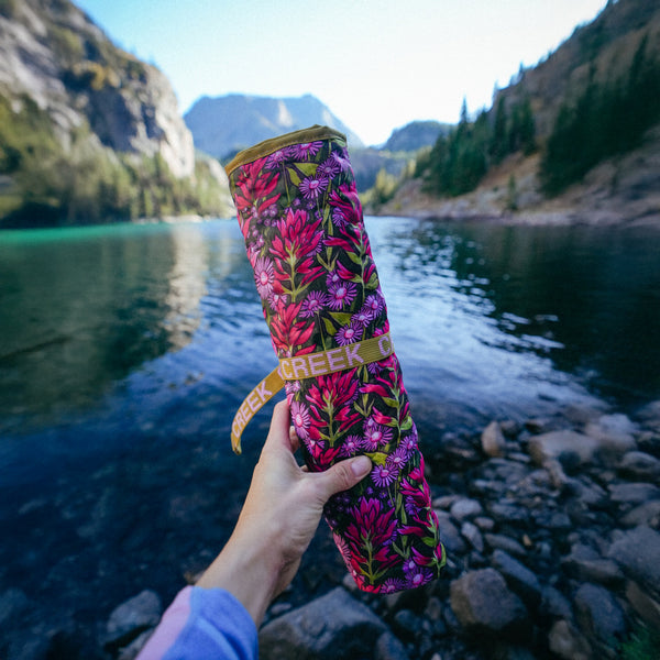 Person holding a floral-patterned Crazy Creek Hex Chair in front of a mountain lake.