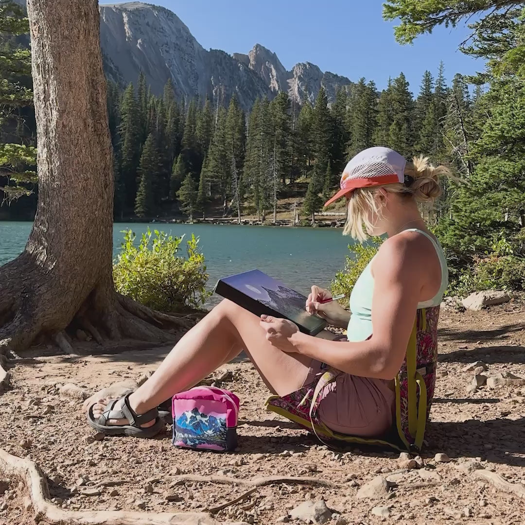 Rachel Pohl sitting next to a blue lake in the mountains painting a landscape