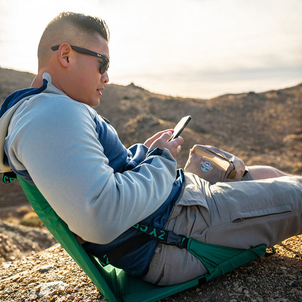 Man sitting in a green chair while looking at his phone on a mountain